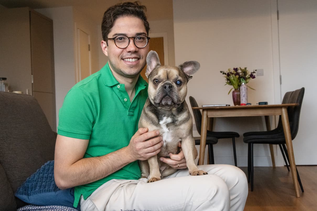 A smiling man sits indoors with a French bulldog resting comfortably on his lap.