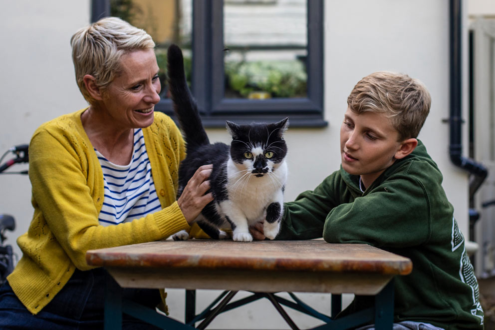 A woman and a boy sitting at a garden table petting a black and white cat.
