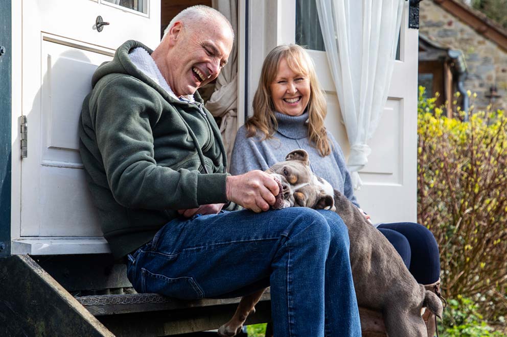 An older couple sitting on wooden steps laughing together while a bulldog rests his head on the man's lap.